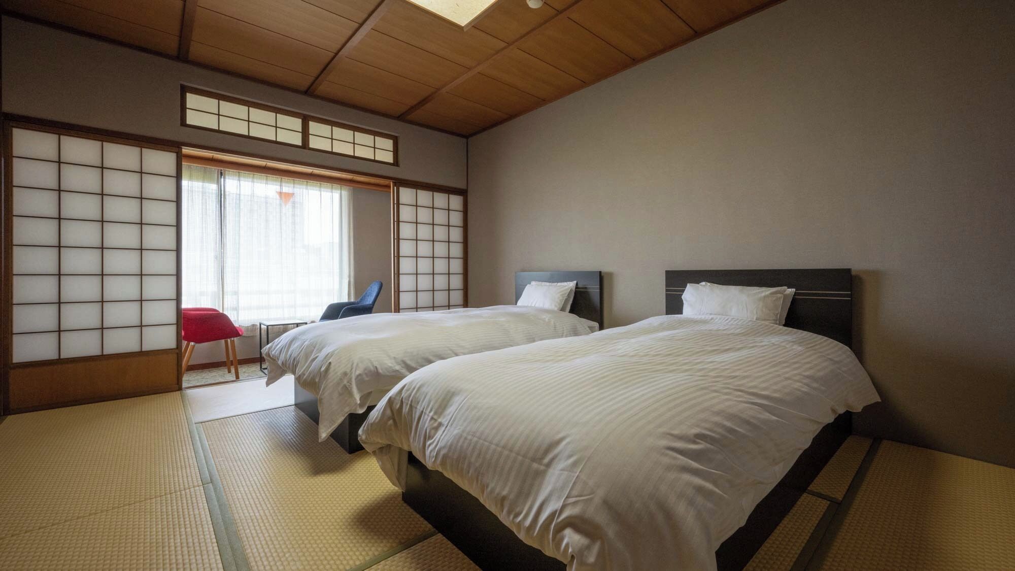 Una habitación tradicional japonesa con suelo de tatami, dos camas con cabeceros oscuros, una silla roja y un panel corredizo shoji.