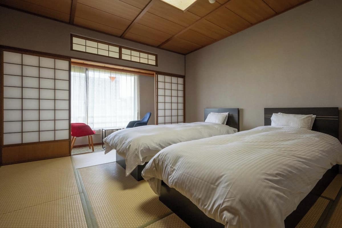 A traditional Japanese-style room with tatami flooring, two beds with dark headboards, a red chair, and a sliding shoji screen.