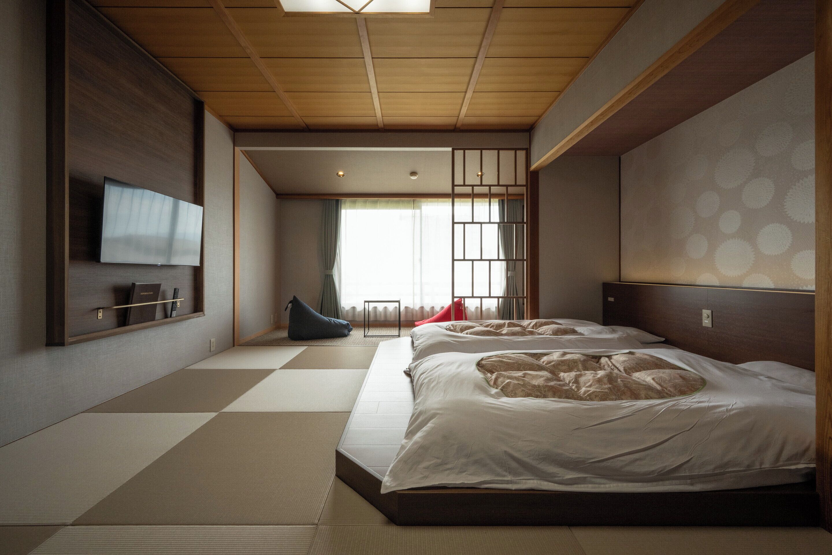 A room with tatami flooring, a bed with white bedding, a television mounted on the wall, and a sliding door with a view of the outside.