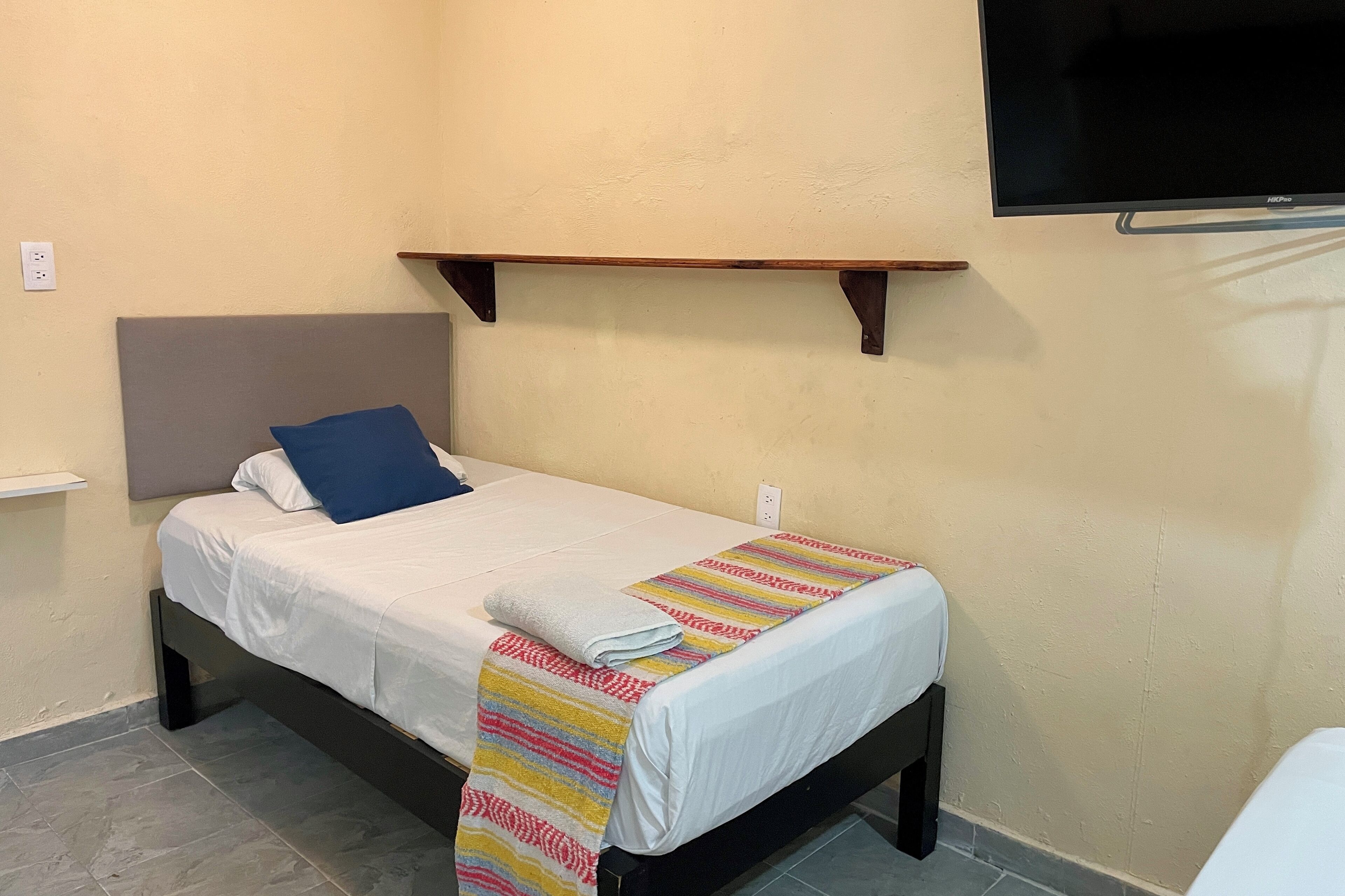 A single bed with a white mattress and a colorful striped blanket, a blue pillow, and a wooden headboard.