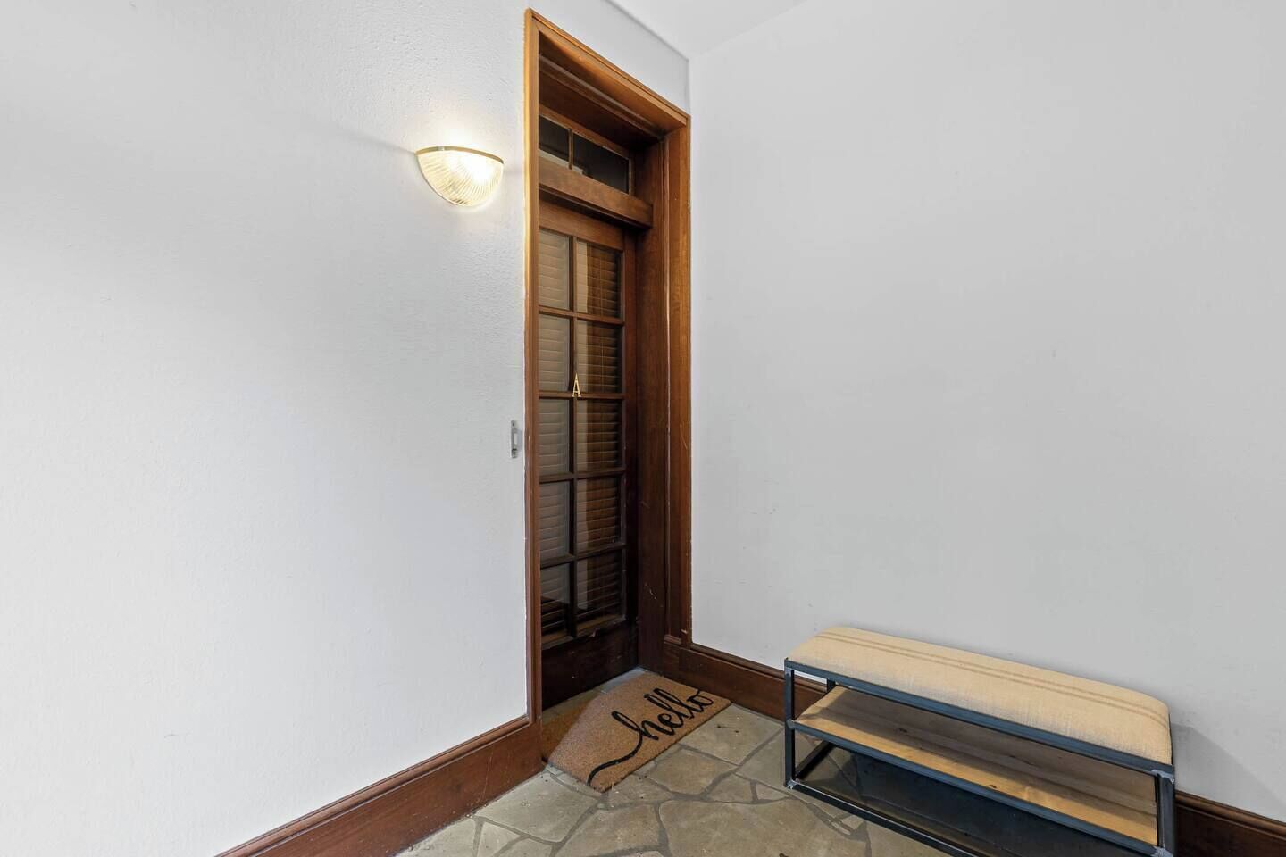 Um corredor com porta de madeira, luminária na parede, tapete de entrada e um banco.