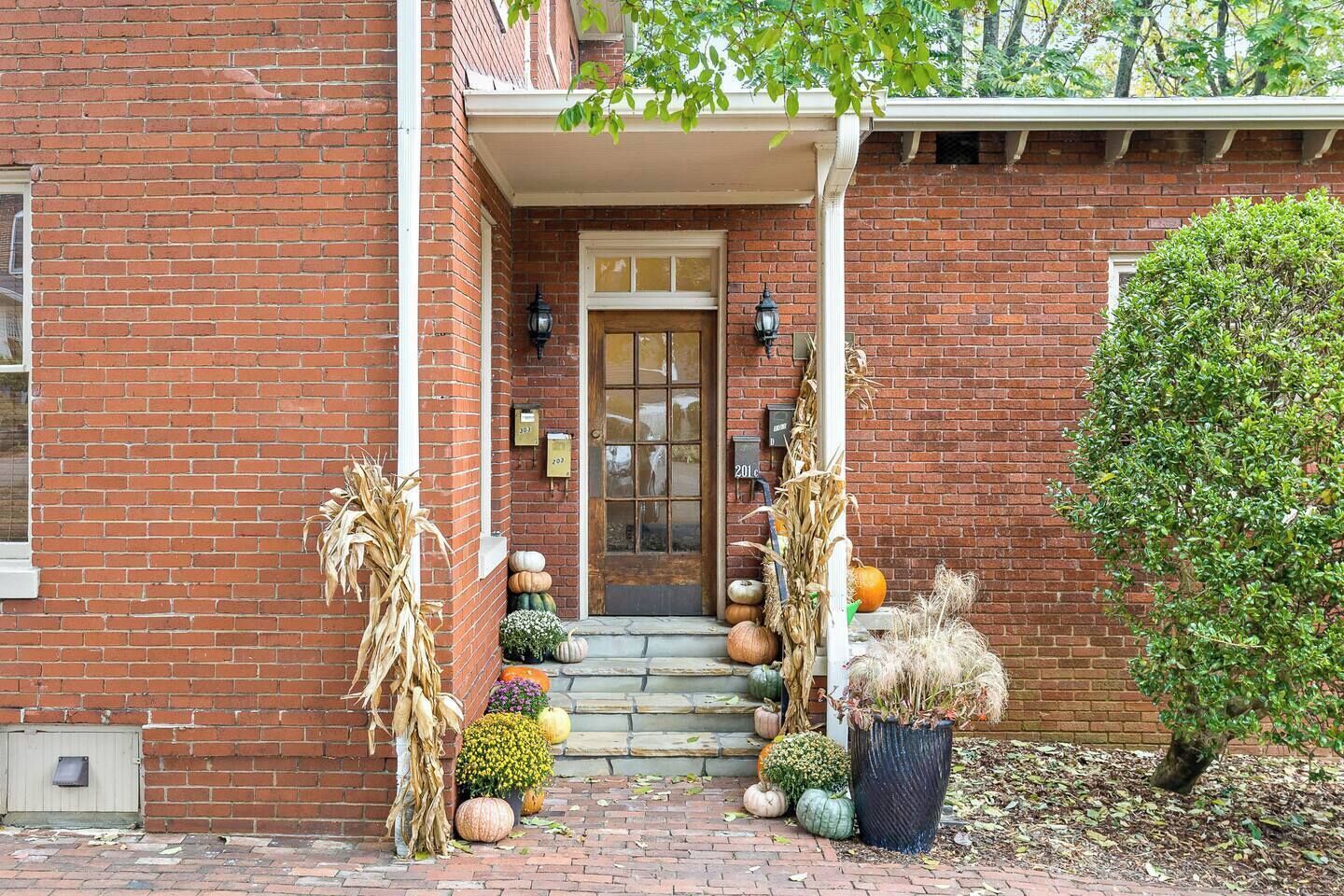 A brick building with a front door, a porch, and a small garden with pumpkins and dried plants.