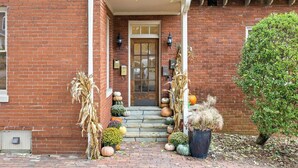 A brick building with a front door, a porch, and a small garden with pumpkins and dried plants.