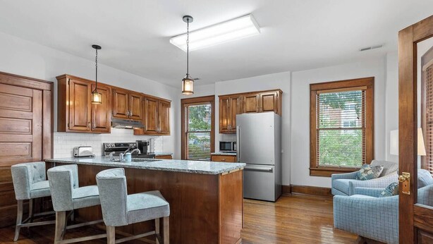 A kitchen with wooden cabinets, a stainless steel refrigerator, a bar area with stools, and a large window with blinds.