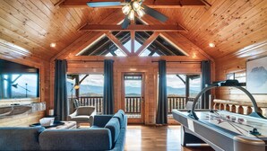 A cozy cabin interior with a wooden ceiling, a sofa, a pool table, and a view of the mountains through large windows.