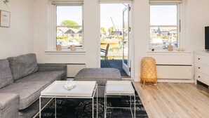 Ein modernes Wohnzimmer mit einem grauen Sofa, einem weißen Couchtisch und einem kleinen Beistelltisch. Drei Fenster bieten Blick auf einen Kanal und Gebäude.