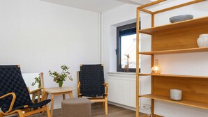 A modern living room with two black chairs, a small round ottoman, a wooden bookshelf, a small table with a vase of flowers, and a window with a view of the outside. - Apartment in Holiday Park- Shared Kitchen (Ulfborg)