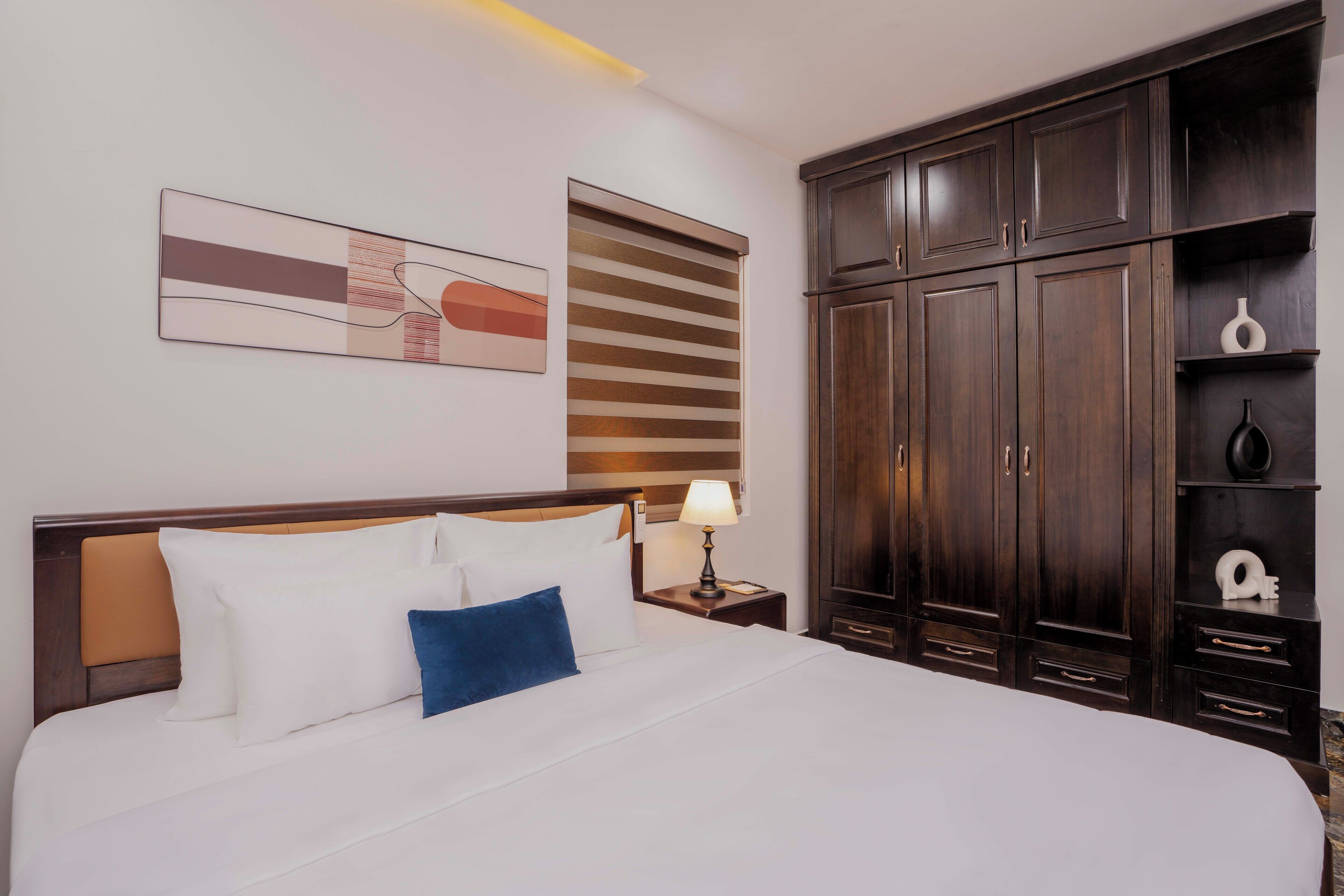 A hotel room with a large bed, a nightstand with a lamp, a wardrobe, and a wall-mounted abstract painting.