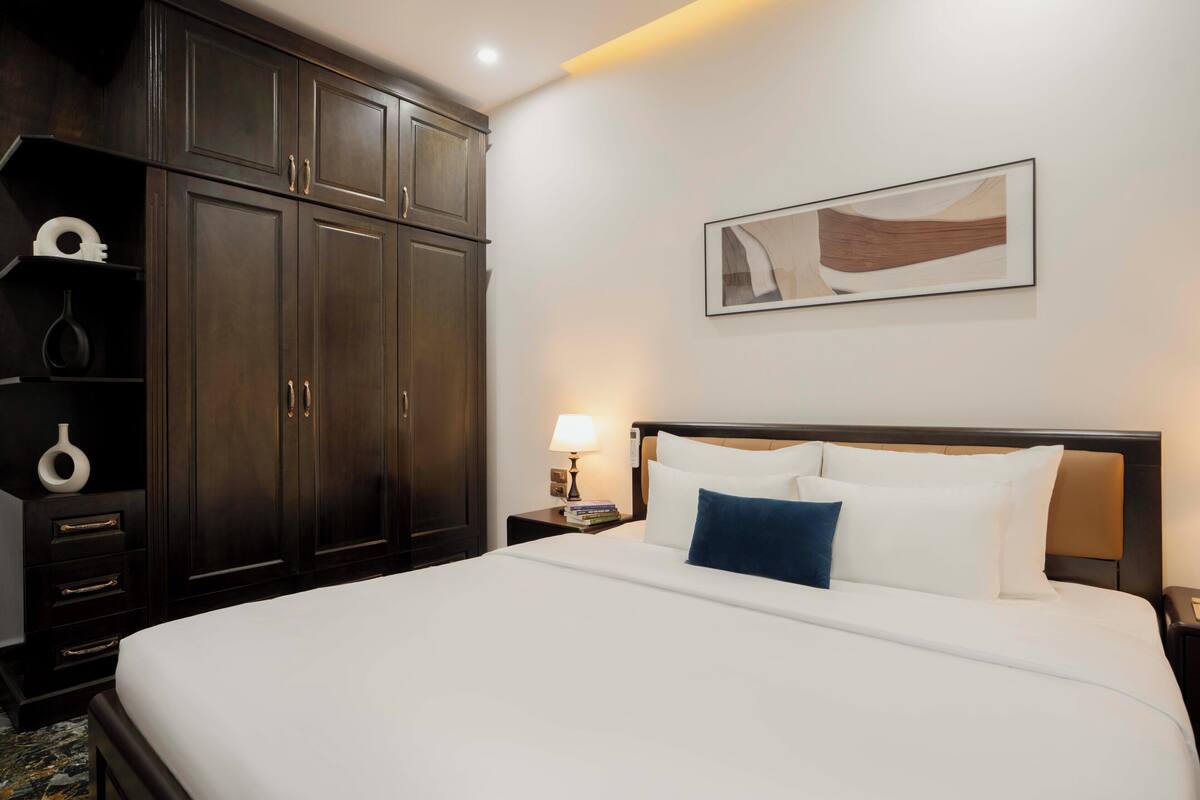 A hotel room with a large bed, dark wooden wardrobe, a nightstand with a lamp, and a framed abstract painting on the wall.