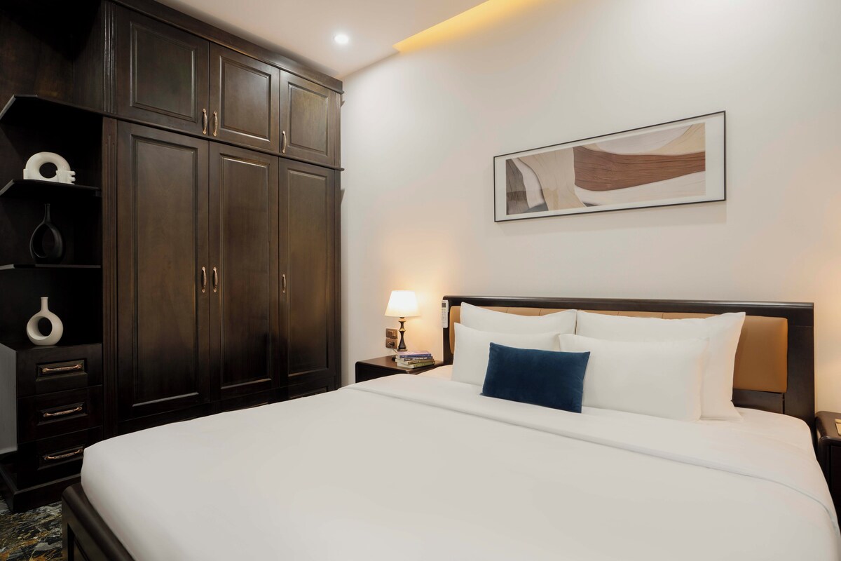 Quarto de hotel com uma cama grande, guarda-roupa de madeira escura, mesa de cabeceira com abajur e um quadro abstrato na parede.