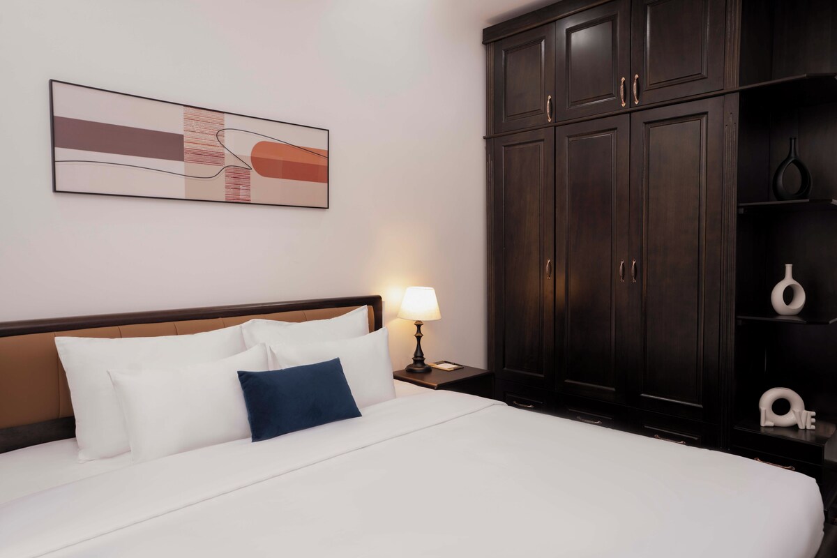 Quarto de hotel com cama grande, mesa de cabeceira com luminária, guarda-roupa de madeira escura e pintura abstrata na parede.