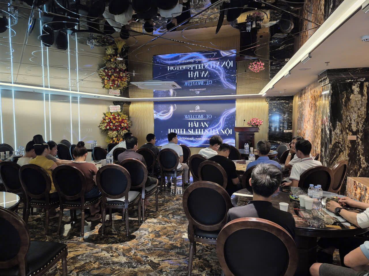 A conference room with round tables and chairs, a large screen displaying a welcome message, and a decorative floral arrangement.