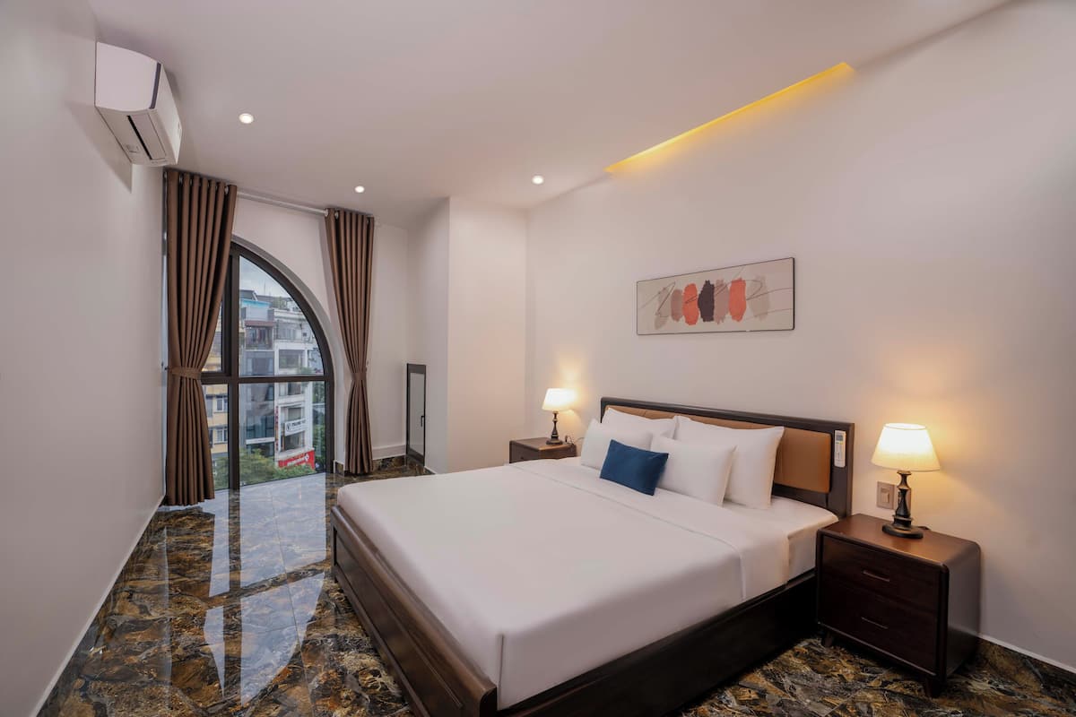 A hotel room with a large bed, two bedside tables with lamps, a window with curtains, and a painting on the wall.
