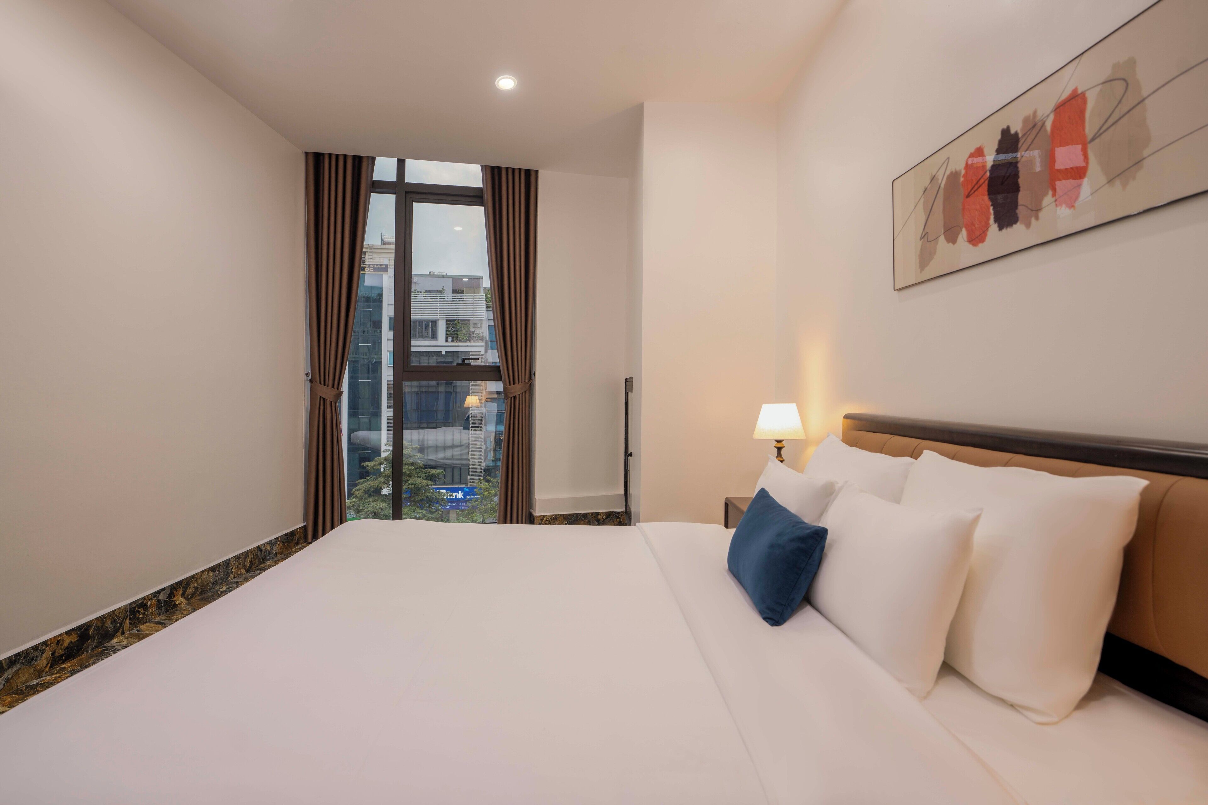 A hotel room with a large bed, a nightstand with a lamp, a window with curtains, and a city view.