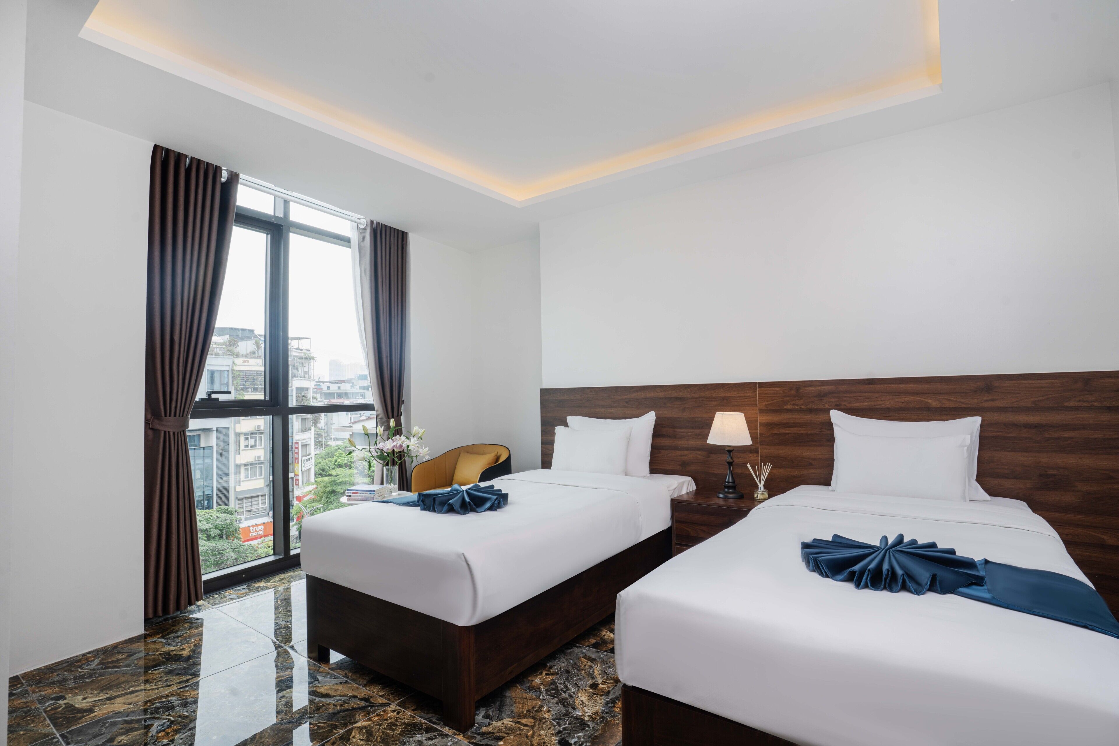 Une chambre d’hôtel avec deux lits, une table de chevet, une lampe, une chaise et une vue sur la ville grâce à de grandes fenêtres.
