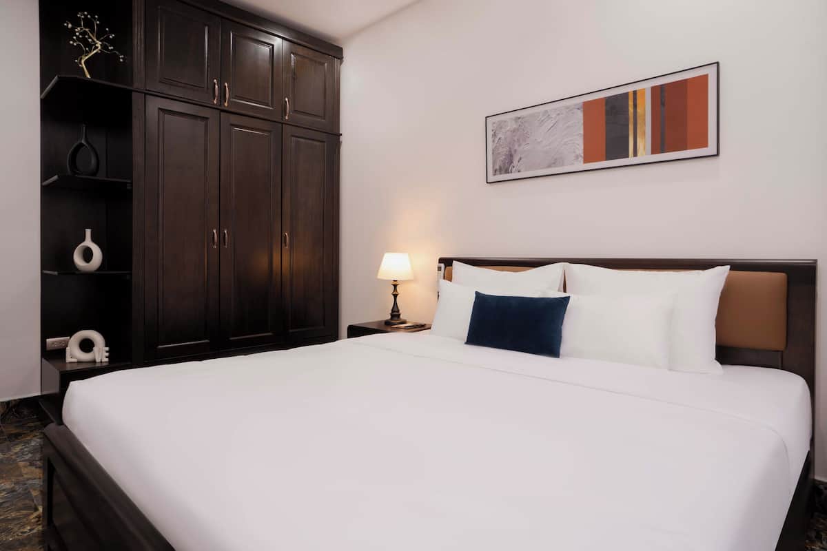 A hotel room with a large bed, dark wooden wardrobe, a bedside table with a lamp, and a framed abstract painting on the wall.