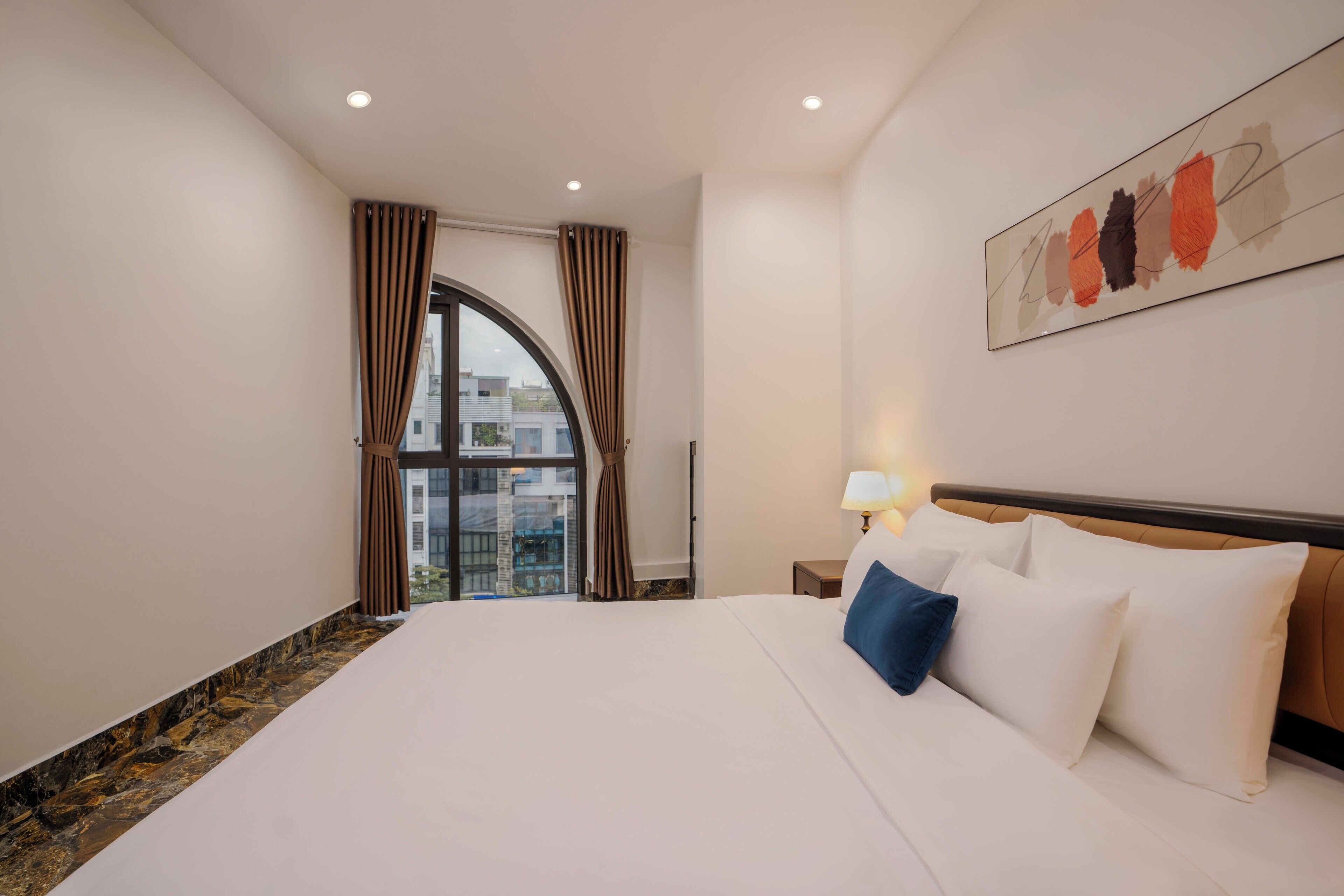 A hotel room with a large bed, a nightstand with a lamp, a view of buildings through a window, and a painting on the wall.
