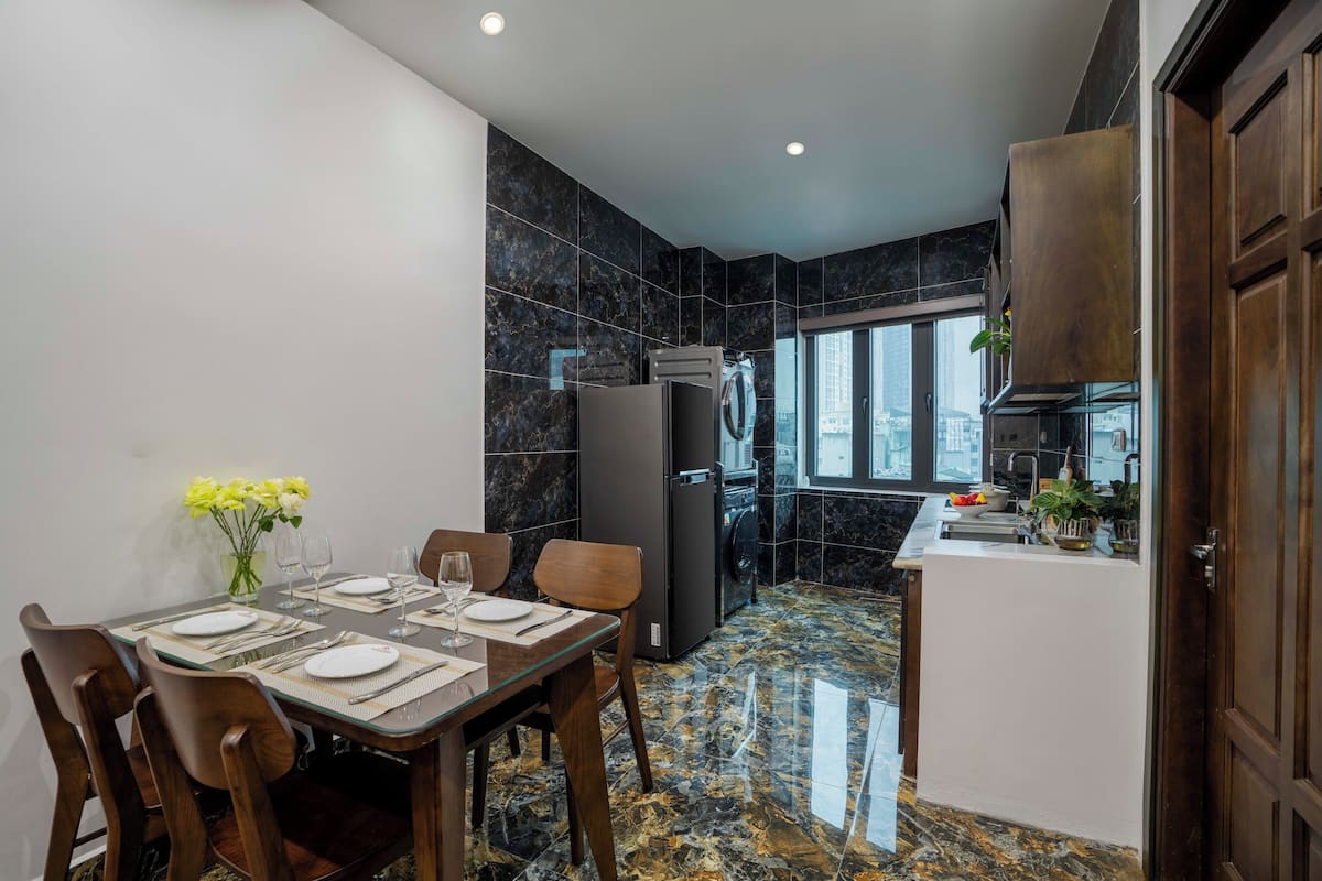 A modern kitchen with dark marble tiles, a dining area with wooden chairs and a glass table, and a view through a large window.