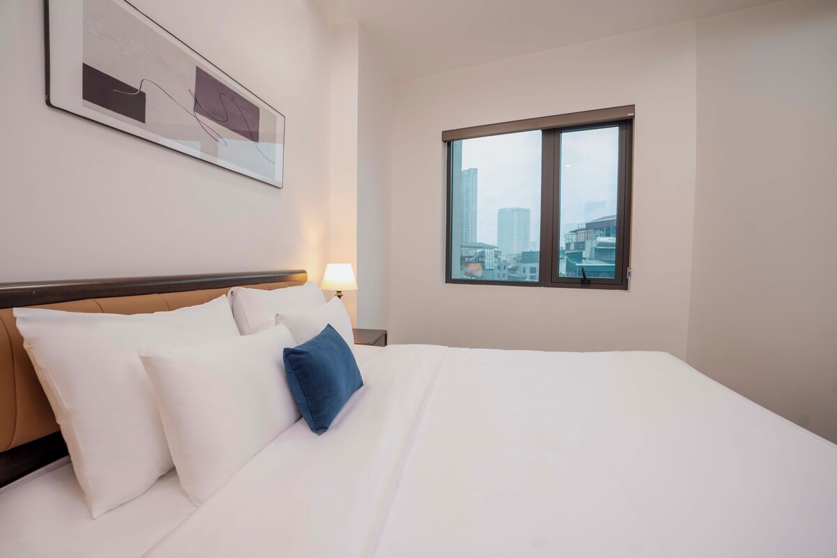 Quarto de hotel com uma cama grande, cabeceira, janela com vista para a cidade e uma obra de arte na parede.