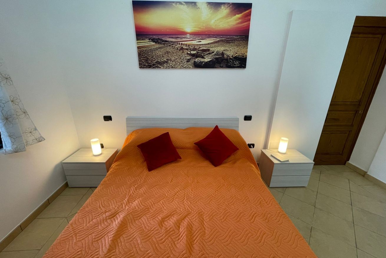 A bedroom with a large bed, two bedside tables with lamps, a nightstand, a wooden door, and a wall-mounted painting.