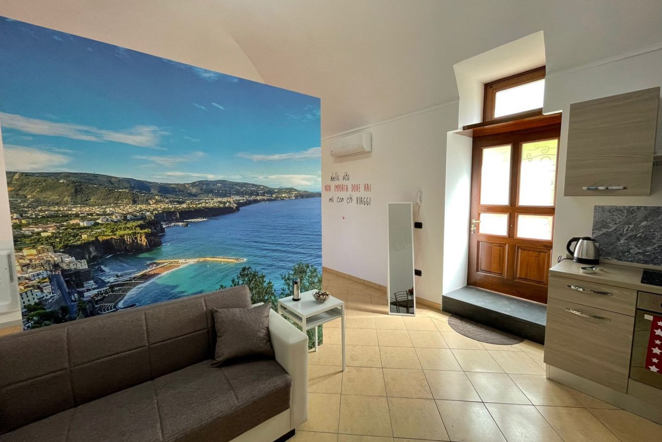 Una sala de estar con un mural grande de una ciudad costera, un sofá, una mesita y una zona de cocina con armarios y una encimera.