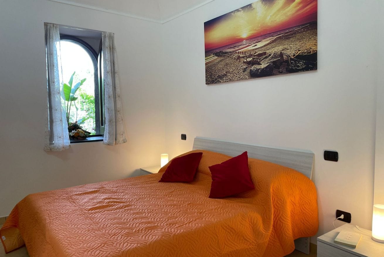 Une chambre à coucher comprenant un lit, des oreillers, une table de chevet, une lampe, une fenêtre avec des rideaux et un tableau accroché au mur.