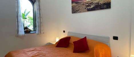 A bedroom with a bed, pillows, a nightstand, a lamp, a window with curtains, and a wall-mounted picture.
