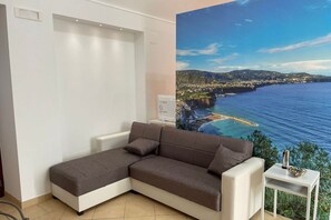 A room with a large window, a white sofa, a small table with a bowl, and a wall-mounted device. - Sweet Sun Studio Apartment – Cozy Comfort Just Minutes from Sorrento ☀️🏡 (Meta)