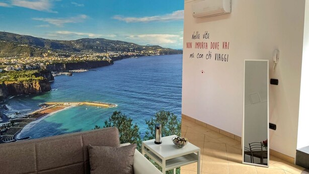 A room with a large window overlooking a coastal landscape, a white wall with a motivational quote, a white table with a bowl and a thermos, and a beige sofa. - Sweet Sun Studio Apartment – Cozy Comfort Just Minutes from Sorrento ☀️🏡 (Meta)
