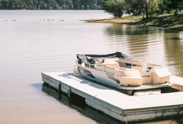 A pontoon boat docked at a wooden pier with a calm lake and trees in the background. - The Lakehouse at Hidden Cove (Fairland)