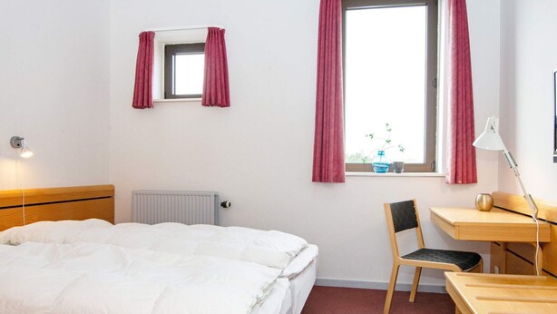 Ein Hotelzimmer mit einem Bett, einem Schreibtisch, einem Stuhl, einem Heizkörper, einem Fenster mit Vorhängen und einer Lampe.