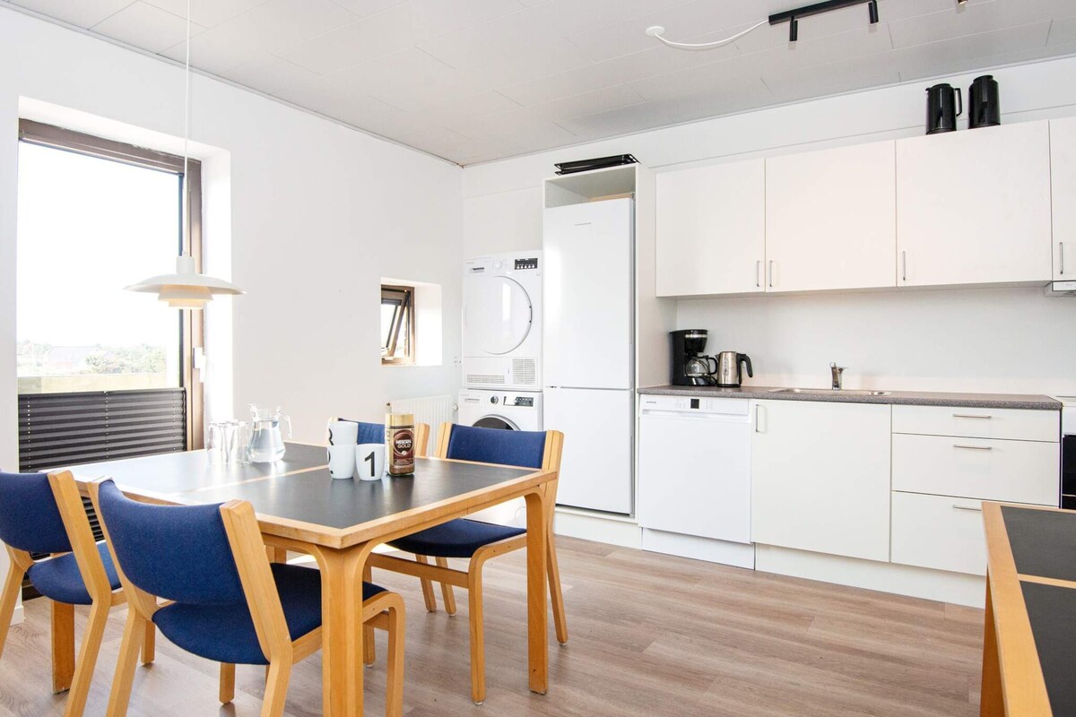 Eine moderne Küche mit weißen Schränken, einem Geschirrspüler und einer Kaffeemaschine. Ein Esstisch mit blauen Stühlen und ein Fenster sorgen für natürliches Licht.