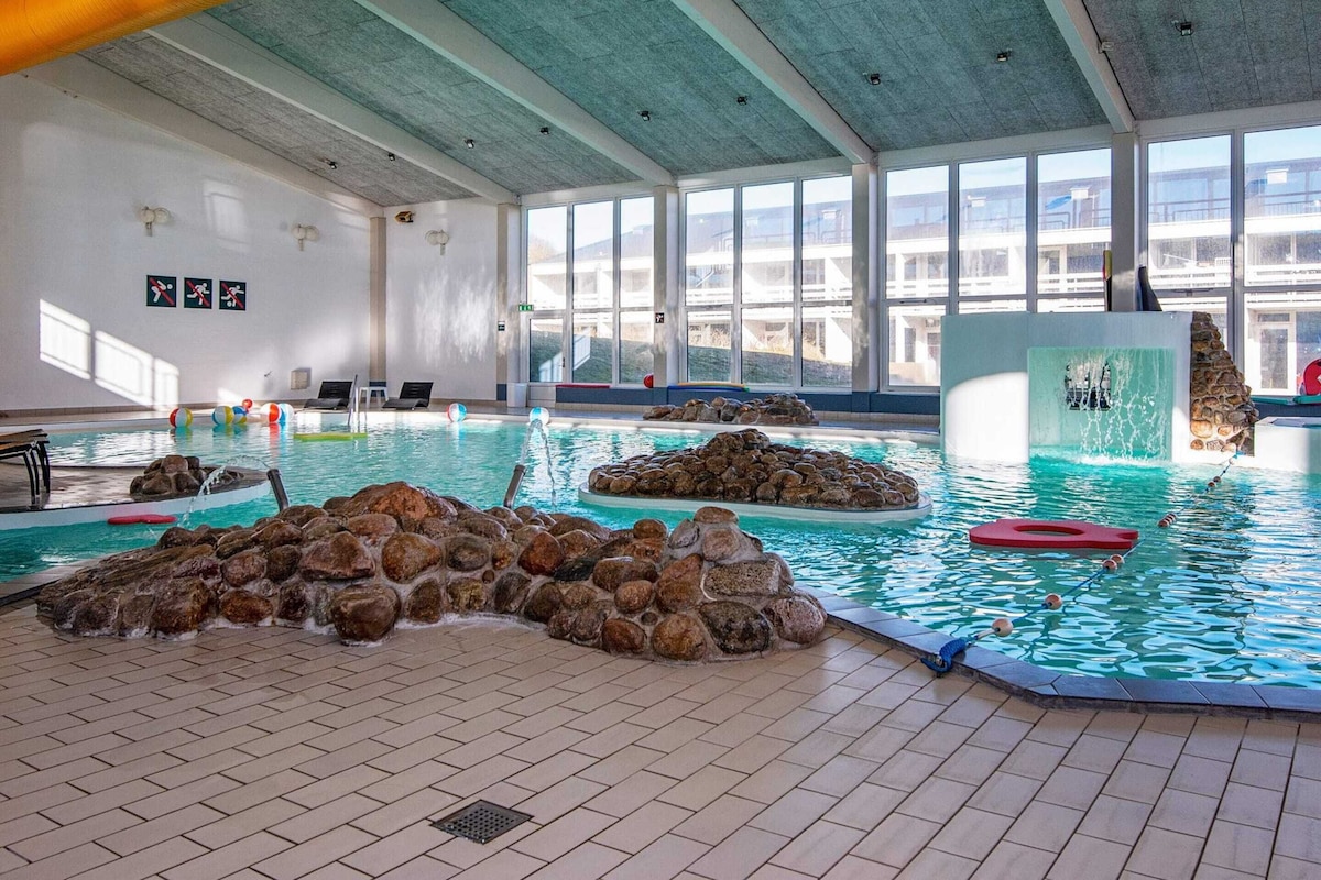 Piscina interna com água cristalina, formações rochosas e vista para o exterior através de grandes janelas.
