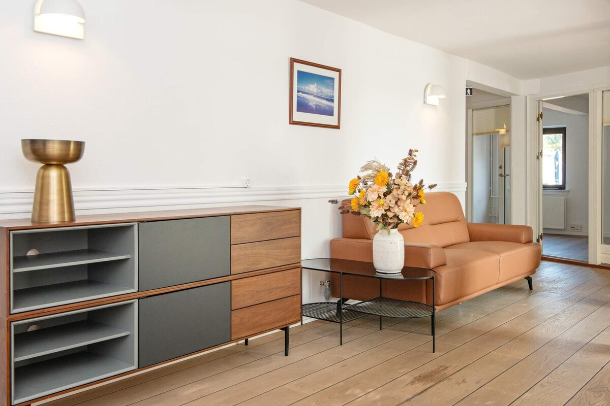 A modern living room with a brown leather sofa, a glass coffee table, a metal sideboard, and a vase with flowers.