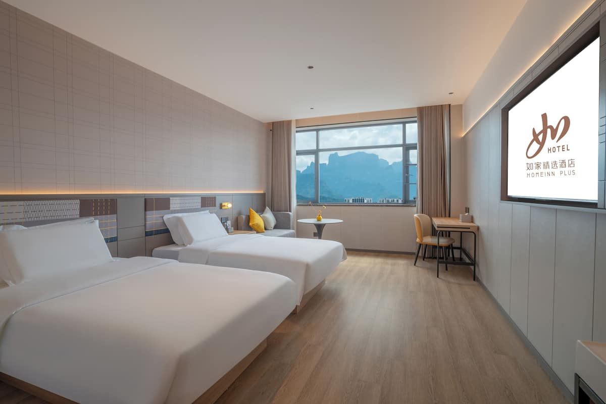 Ein Hotelzimmer mit zwei Betten, einem kleinen Tisch, einem Stuhl und einem großen Fenster mit Blick auf die Berge.