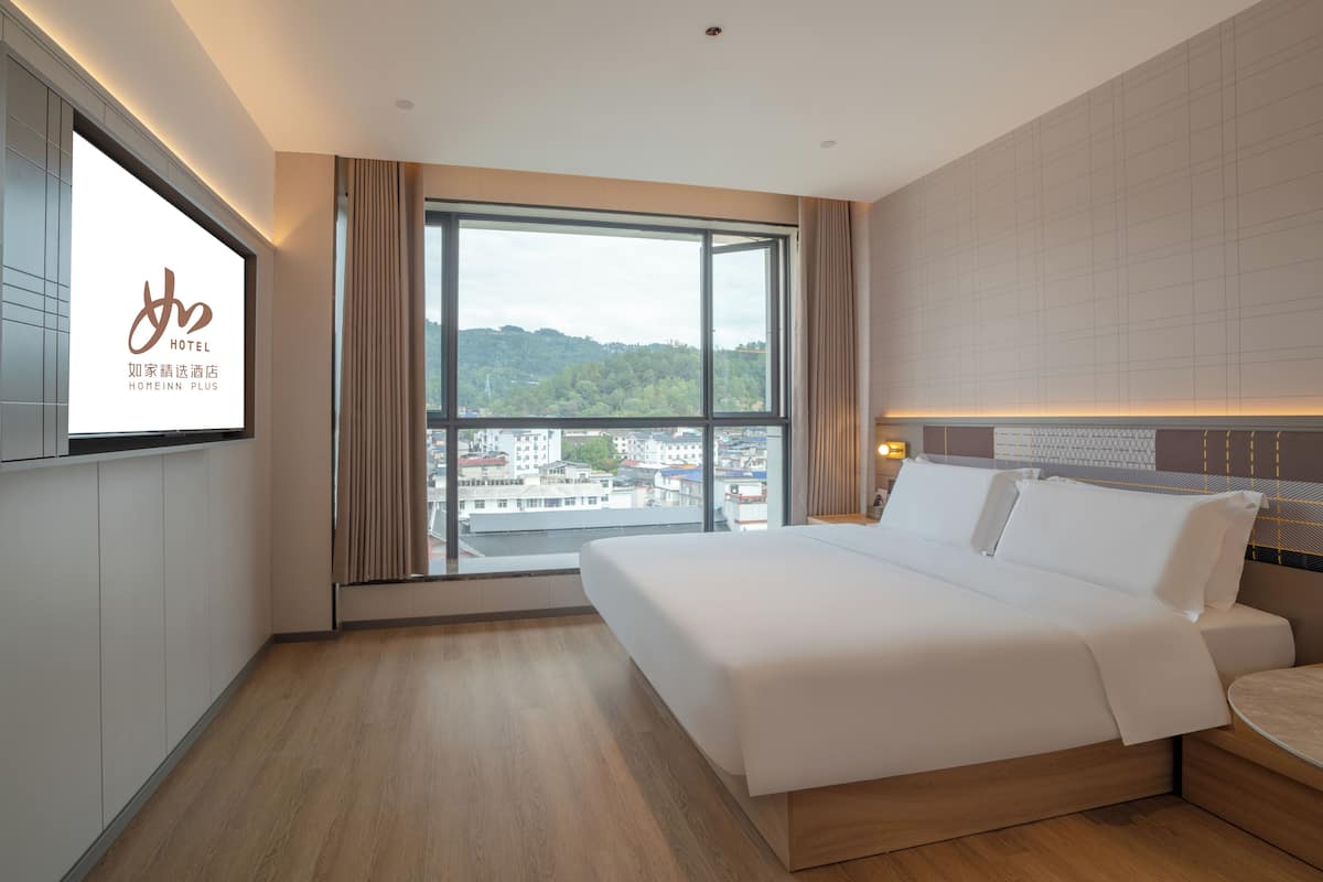 Ein Hotelzimmer mit großem Fenster, einem Bett, einem Fernseher und Blick auf Gebäude und Grünflächen.
