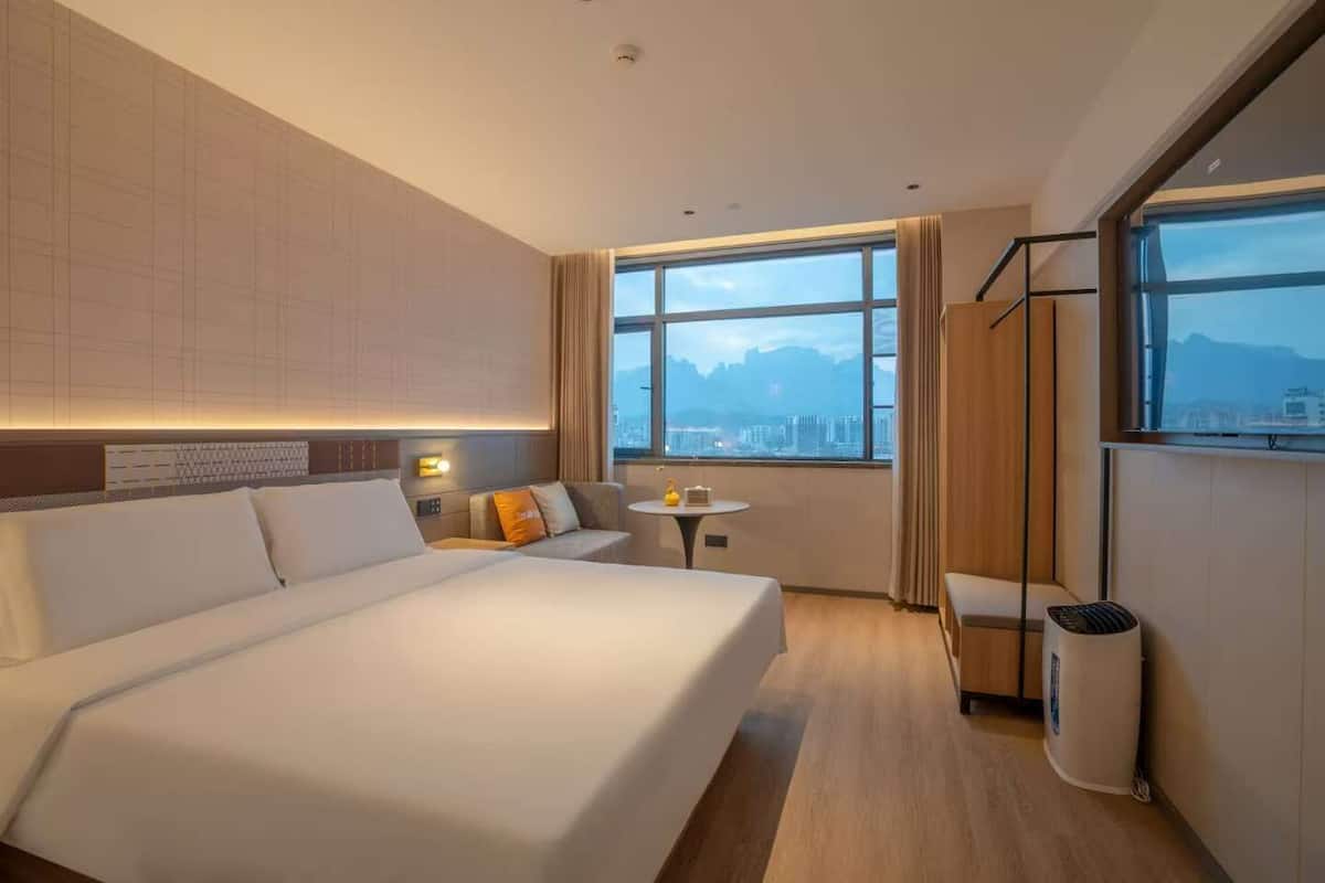 Ein modernes Hotelzimmer mit einem großen Bett, einem kleinen Tisch und Blick auf die Stadt durch große Fenster.