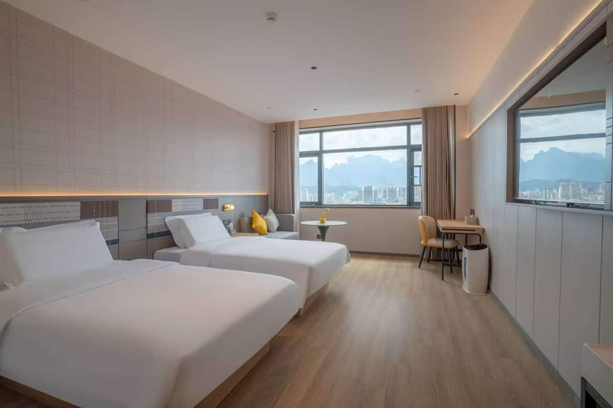 Ein Hotelzimmer mit zwei Betten, einem kleinen Tisch, einem Sessel und Blick auf die Stadt durch große Fenster.