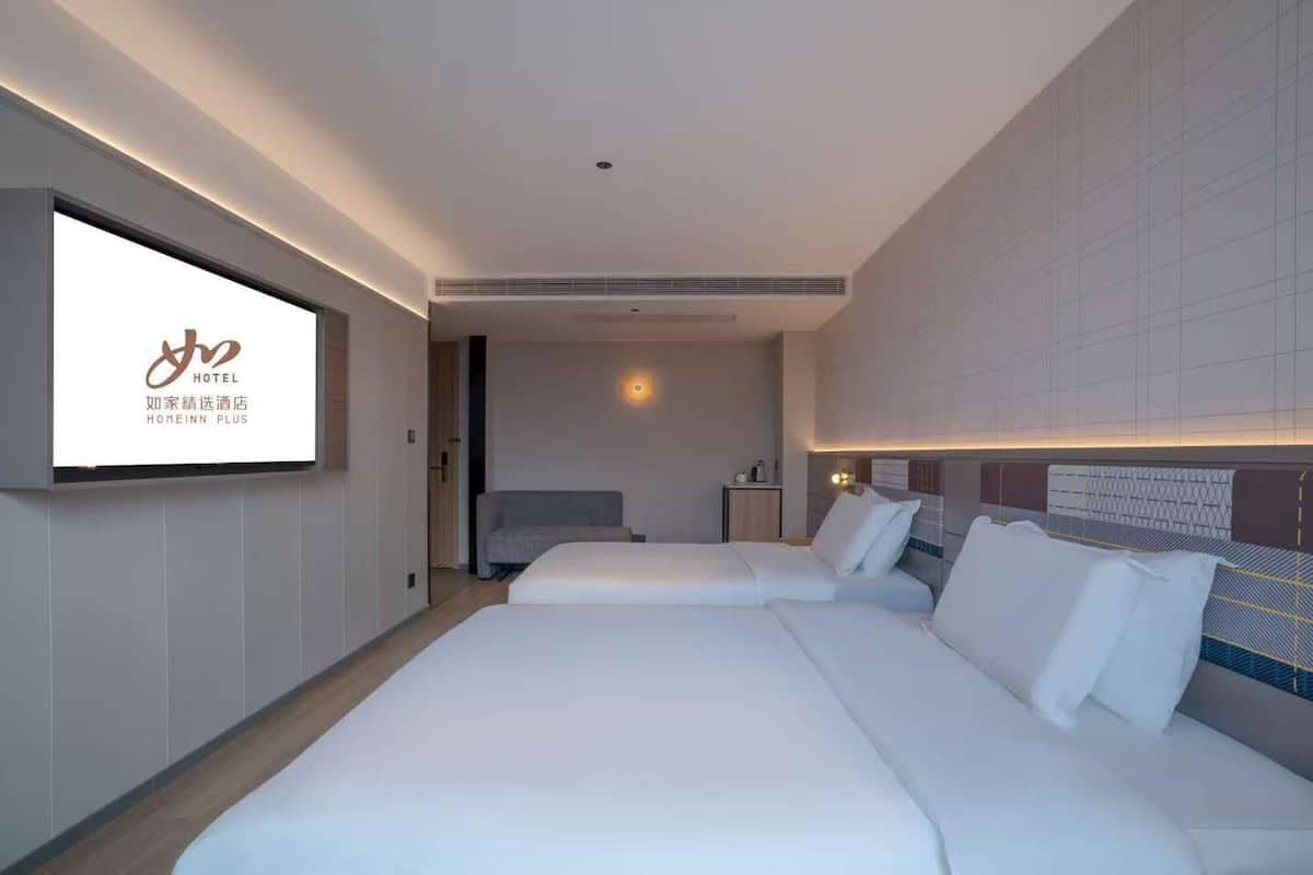 Ein Hotelzimmer mit zwei Betten, einem Fernseher der die Marke des Hotels zeigt, und einem modernen Design mit einem gemusterten Wand- und Deckenbereich.