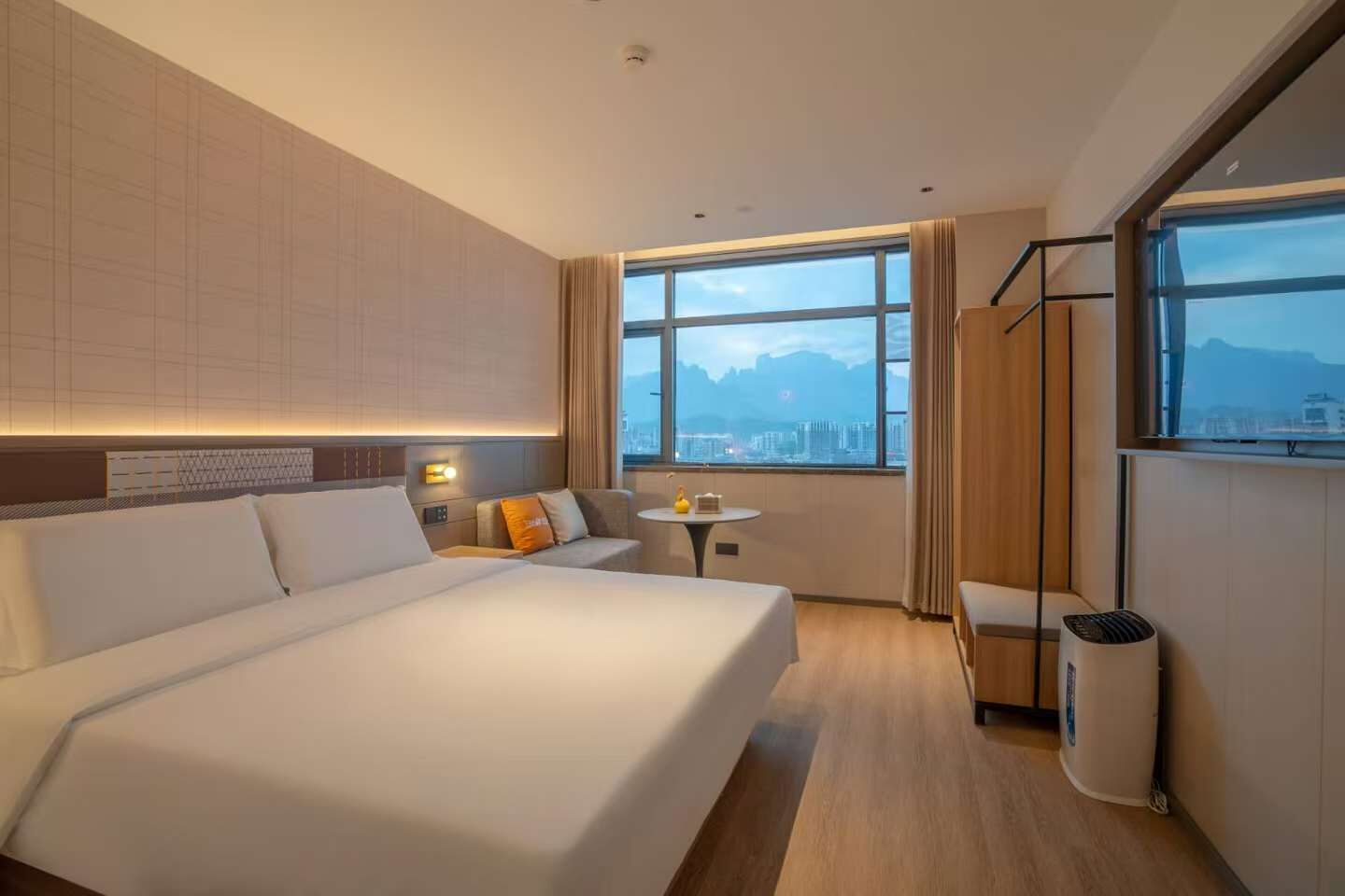 Quarto de hotel moderno com uma cama grande, uma mesa pequena e vista para a cidade por grandes janelas.