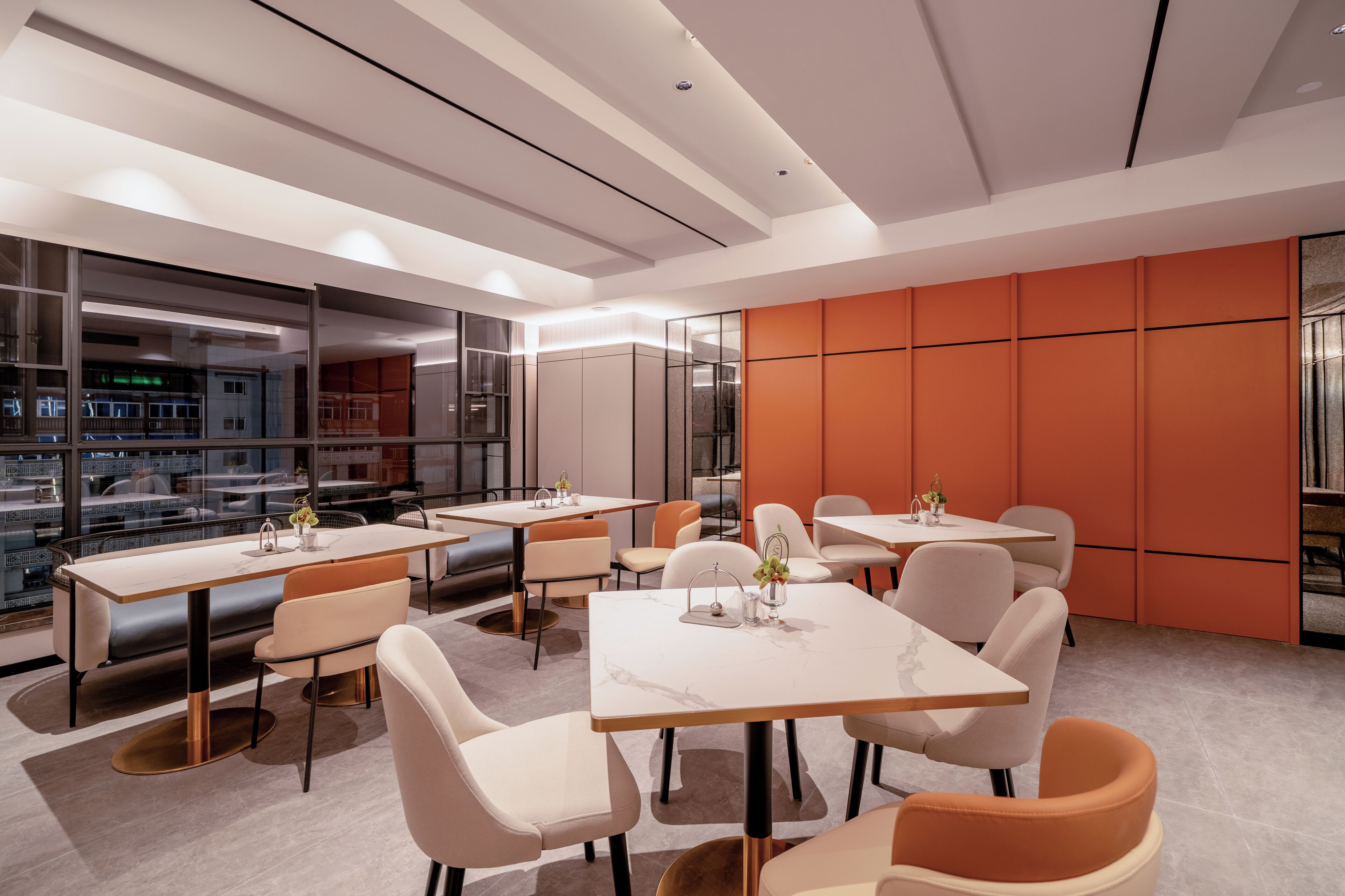 A modern dining area with tables and chairs, large windows, and orange accent walls.