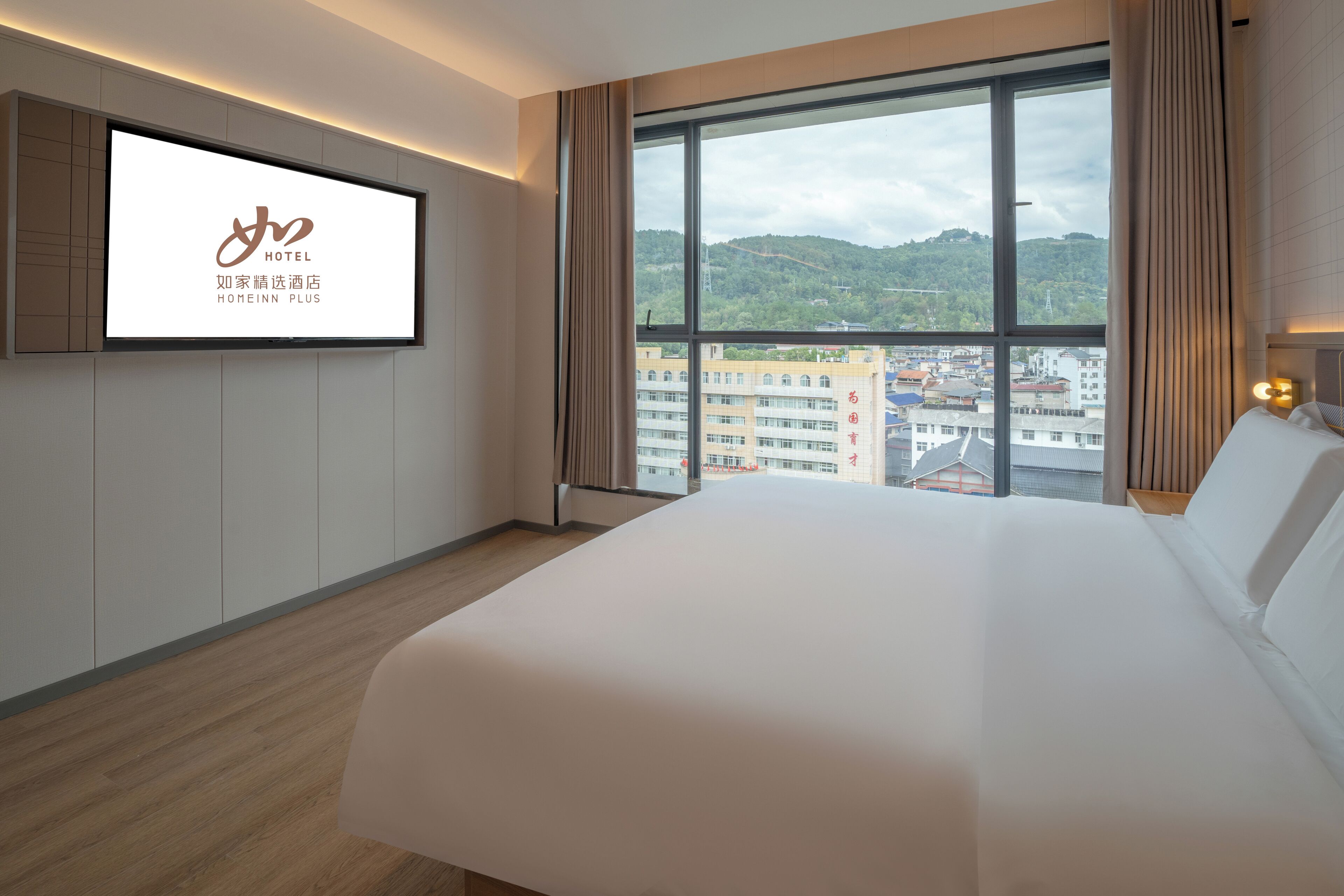 Ein Hotelzimmer mit einem großen Bett, einem an der Wand befestigten Fernseher und Blick auf eine Stadt durch große Fenster.