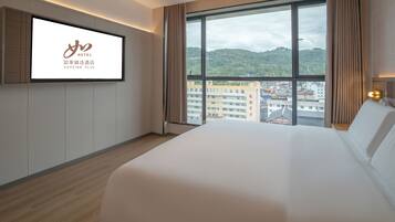 A hotel room with a large bed, a TV mounted on the wall, and a view of a cityscape through large windows.