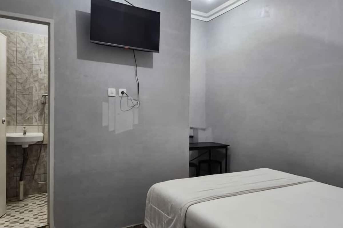 A hotel room with a bed, a TV mounted on the wall, a desk, and a bathroom with a sink and mirror.