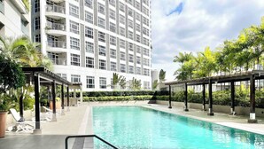 A swimming pool with a clear blue water, surrounded by lounge chairs and a covered area, with a tall building in the background. - Comfy condo. Best location on Shaw Blvd, front of Shang Mall. Short walk to SM. (Mandaluyong City)