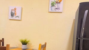 Un comedor con una mesa de madera, sillas y una planta en maceta. Hay dos cuadros colgados en la pared que representan utensilios de cocina. Se ve un refrigerador negro a la derecha.