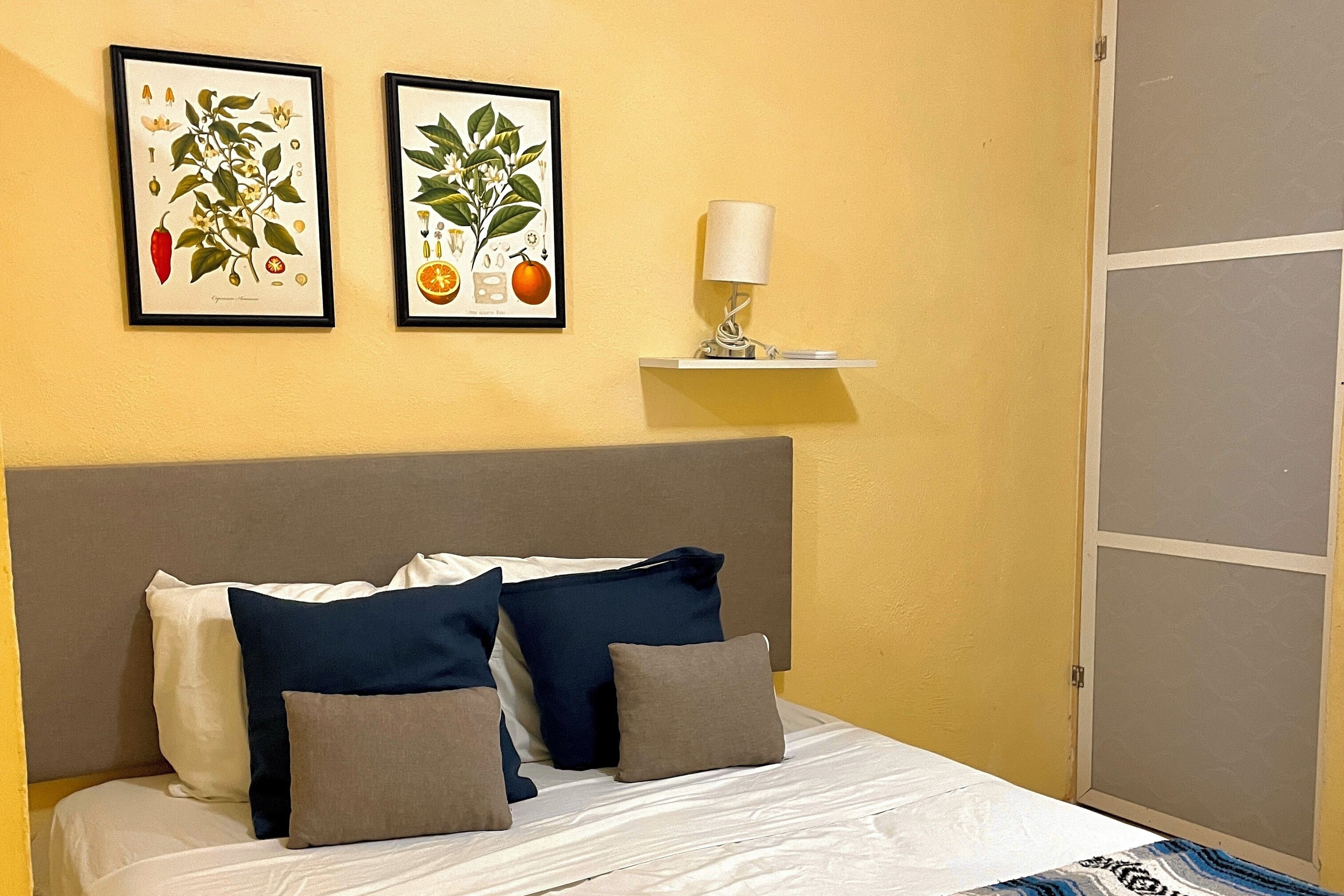 Una cama bien hecha con cabecera gris, ropa de cama blanca y múltiples almohadas en azul marino y gris. Dos láminas botánicas enmarcadas cuelgan de la pared sobre la cama.