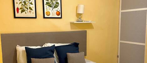 A neatly made bed with a grey headboard, white bedding, and multiple pillows in navy and grey. Two framed botanical prints are hung on the wall above the bed.