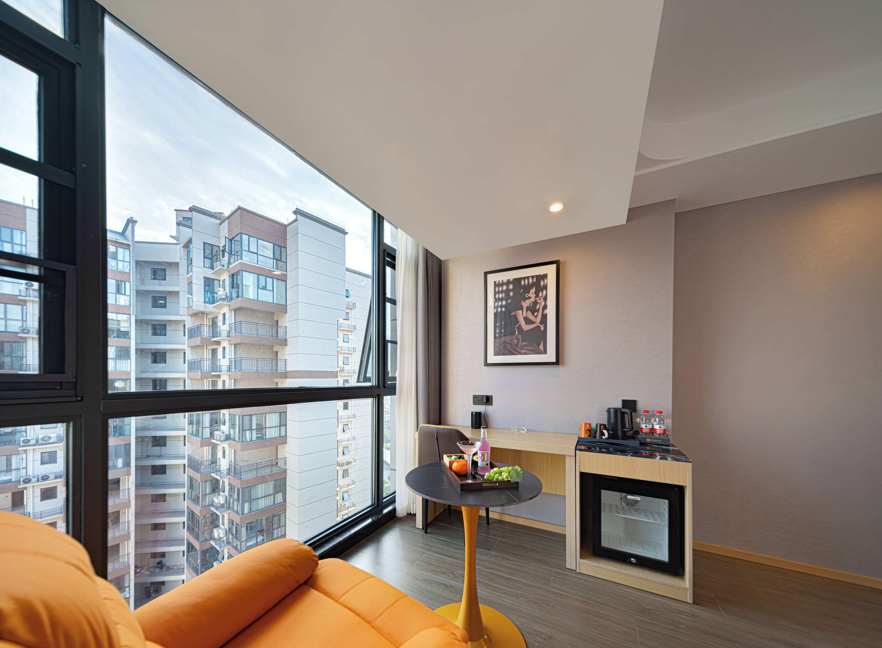 A modern apartment with a large window, a small round table with a tray of fruit, a sofa, and a kitchenette.
