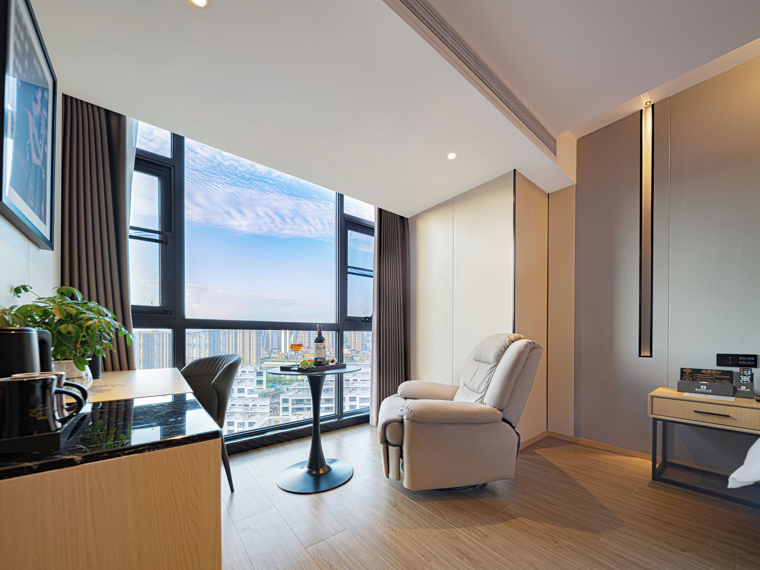 A modern hotel room with a large window, a dining table, a sofa, and a small desk.